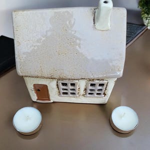 Puede incluir: Un portavelas de cerámica con forma de casa con un esmalte blanco. La casa tiene dos ventanas y una puerta. Hay tres velas de té sobre una superficie marrón.