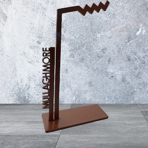 Puede incluir: Escultura de metal marrón con la palabra "MULLAGHMORE" recortada verticalmente. La escultura tiene una base rectangular, soportes verticales y una parte superior en zigzag. El fondo es gris y blanco texturizado.