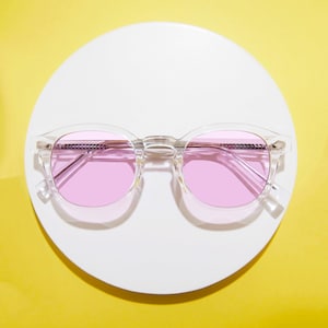May include: Clear-framed sunglasses with pink lenses. The round frames have a classic design, and the lenses are a vibrant pink color. The sunglasses are set against a white circular background on a yellow surface.