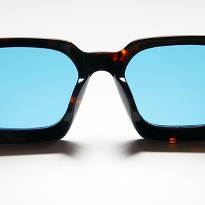 Puede incluir: Un par de gafas de sol de carey con lentes azules. Las gafas tienen una forma cuadrada y est&aacute;n hechas de pl&aacute;stico.