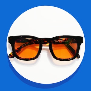 May include: Square-framed sunglasses with a tortoiseshell pattern and orange lenses. The glasses are set against a white circular background with a blue backdrop. The frame has a classic, bold design.