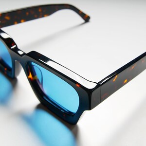 Puede incluir: Gafas de sol con montura de carey y lentes azules. Las gafas tienen una forma cuadrada y una montura gruesa.