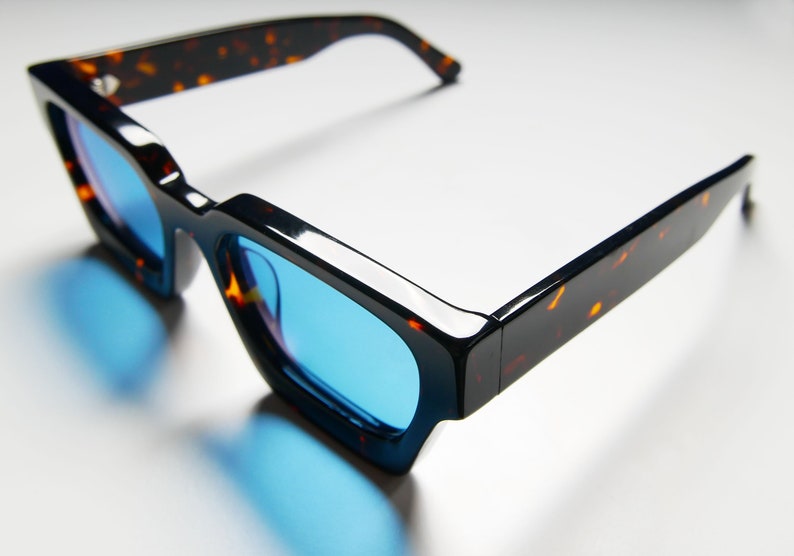 Puede incluir: Gafas de sol con montura de carey y lentes azules. Las gafas tienen una forma cuadrada y una montura gruesa.