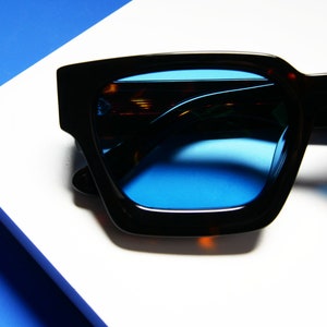 Puede incluir: Gafas de sol cuadradas de color negro y marr&oacute;n con lentes azules. Las gafas de sol est&aacute;n sobre una superficie blanca con un fondo azul.