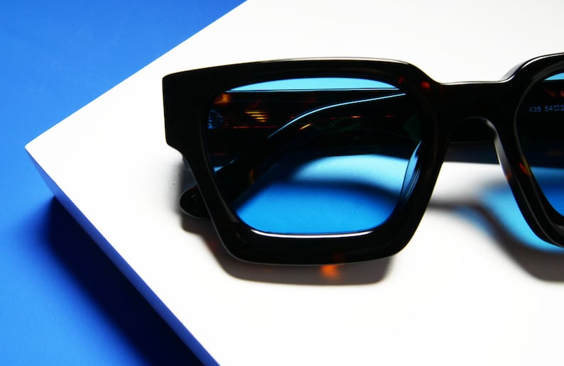 Puede incluir: Gafas de sol cuadradas de color negro y marr&oacute;n con lentes azules. Las gafas de sol est&aacute;n sobre una superficie blanca con un fondo azul.