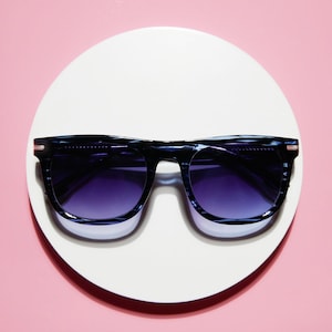 May include: Sunglasses with a dark blue frame and gradient purple lenses rest on a white circular surface against a pink background. The sunglasses have a classic rectangular shape with a subtle pattern.