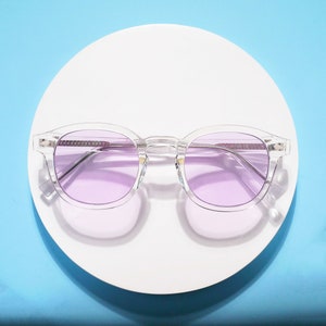 Può includere: Occhiali da sole con montatura in plastica trasparente e lenti viola. Gli occhiali da sole sono su un piatto bianco.