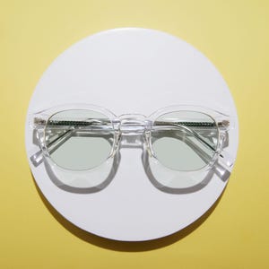 May include: Clear-framed eyeglasses with light green lenses rest on a white circular surface against a yellow background. The glasses have a classic round shape with a keyhole bridge and silver-toned metal accents on the temples.