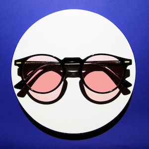 May include: Black-framed sunglasses with pink lenses rest on a white circular surface against a blue background. The sunglasses have a classic round shape with a keyhole bridge and small gold accents on the temples. The pink lenses provide a soft, warm hue.
