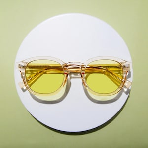 May include: Clear-framed sunglasses with bright yellow lenses. The round lenses are set in a transparent frame with a keyhole bridge. The sunglasses are displayed on a white circular surface against a light green background.