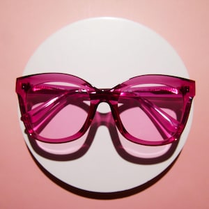 May include: A pair of oversized, pink-tinted sunglasses with a thick, rectangular frame. The glasses are set against a white circular backdrop, with a soft pink background. The lenses and frame are a matching shade of pink.