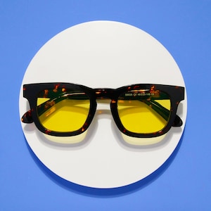 May include: Square-framed sunglasses with a tortoiseshell pattern and yellow lenses. The glasses are set against a white circular background on a blue surface. The frame has a dark brown and amber pattern.