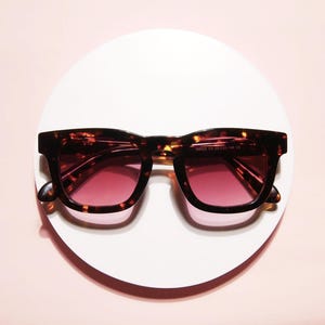 May include: Square-framed sunglasses with a tortoiseshell pattern and gradient pink lenses. The sunglasses are displayed on a white circular surface against a pink background. The frame has a classic, bold design.