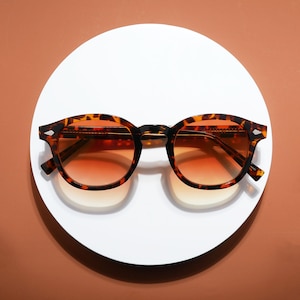 May include: Round sunglasses with a tortoiseshell pattern frame and gradient orange lenses. The sunglasses are displayed on a white circular surface against a brown background. The frames have a classic design with a modern twist.
