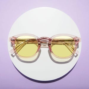 May include: Pink-framed sunglasses with yellow lenses rest on a white circular surface against a purple background. The glasses have a classic round shape with a keyhole bridge and gold-colored arms.