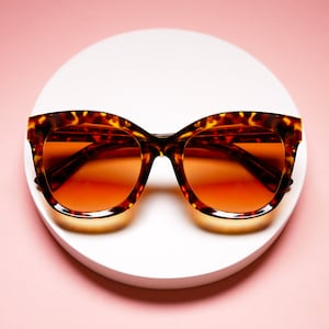 May include: Tortoiseshell sunglasses with orange gradient lenses. The frames have a classic cat-eye shape and are set against a white circular backdrop with a pink background. The sunglasses are a stylish accessory.