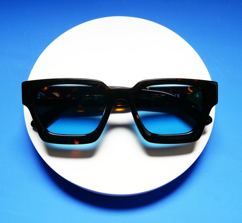 Puede incluir: Gafas de sol de carey con lentes azules. Las gafas tienen una forma cuadrada y est&aacute;n sobre una superficie blanca.