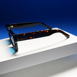 Puede incluir: Gafas de sol de carey con montura negra y lentes azules. Las gafas de sol est&aacute;n sobre una superficie blanca.