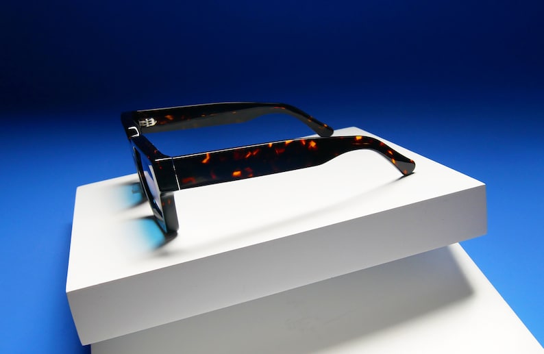 Puede incluir: Gafas de sol de carey con montura negra y lentes azules. Las gafas de sol est&aacute;n sobre una superficie blanca.