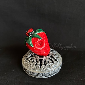 May include: A red and green ceramic strawberry with a ladybug on top, sitting on a silver decorative base.