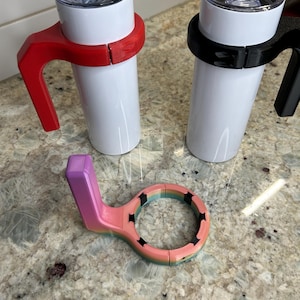 May include: Two white insulated tumblers with clear lids, one with a red handle and the other with a black handle. A multi-colored tumbler handle is also shown, with a purple handle and a ring with black star cutouts.