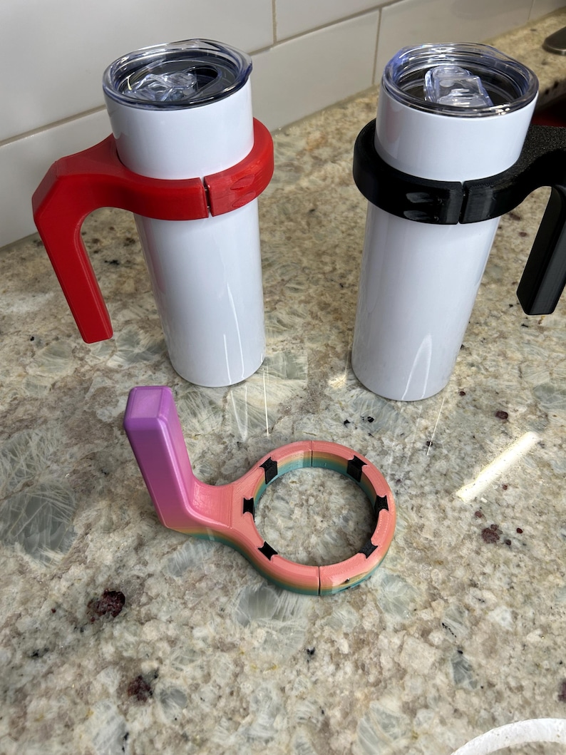 May include: Two white insulated tumblers with clear lids, one with a red handle and the other with a black handle. A multi-colored tumbler handle is also shown, with a purple handle and a ring with black star cutouts.