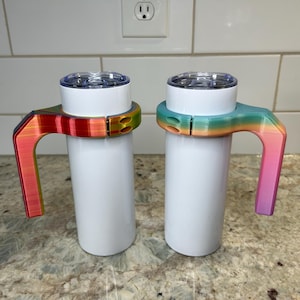 Può includere: Due bicchieri isolanti bianchi con coperchi trasparenti. Ogni bicchiere ha un manico colorato con sfumatura arcobaleno. Un manico passa dal rosso al giallo al verde, l'altro dal verde acqua all'arancione al rosa. I manici sono attaccati ai bicchieri.