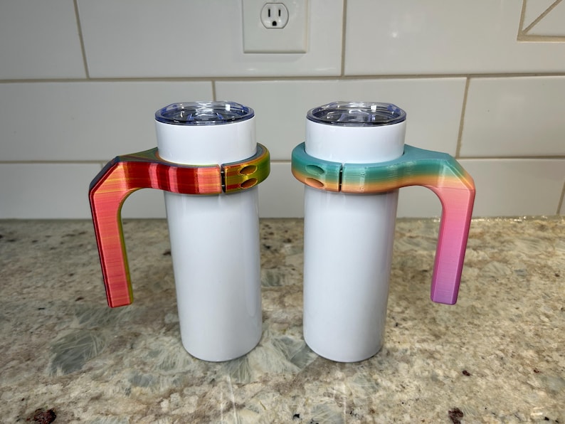 May include: Two white insulated tumblers with clear lids. Each tumbler has a colorful, rainbow-gradient handle. One handle transitions from red to yellow to green, the other from teal to orange to pink. The handles are attached to the tumblers.