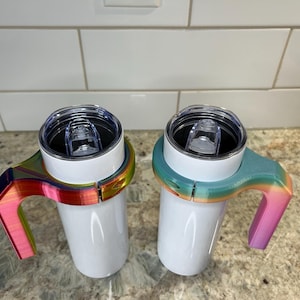 May include: Two white insulated tumblers with clear lids, each fitted with a colorful, rainbow-hued handle. The handles are designed to fit snugly around the tumblers, providing a comfortable grip. The tumblers are designed to hold approximately 10-12 ounces of liquid.