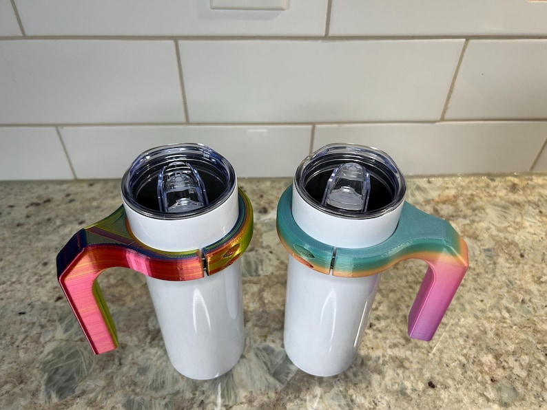May include: Two white insulated tumblers with clear lids, each fitted with a colorful, rainbow-hued handle. The handles are designed to fit snugly around the tumblers, providing a comfortable grip. The tumblers are designed to hold approximately 10-12 ounces of liquid.