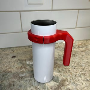 May include: A white travel mug with a red handle. The mug has a silver interior and is held by a red plastic handle that wraps around the middle. The handle is designed for easy carrying and use. The mug is cylindrical and appears to be made of metal.