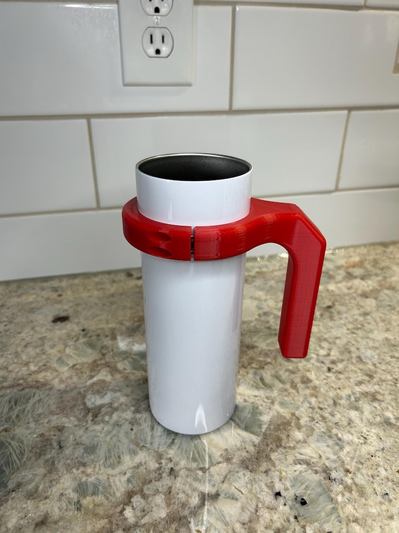May include: A white travel mug with a red handle. The mug has a silver interior and is held by a red plastic handle that wraps around the middle. The handle is designed for easy carrying and use. The mug is cylindrical and appears to be made of metal.