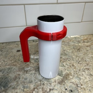 May include: A white tumbler with a red handle. The handle is attached to the tumbler with a clasp. The tumbler is sitting on a granite countertop. The tumbler is designed to hold hot or cold beverages.