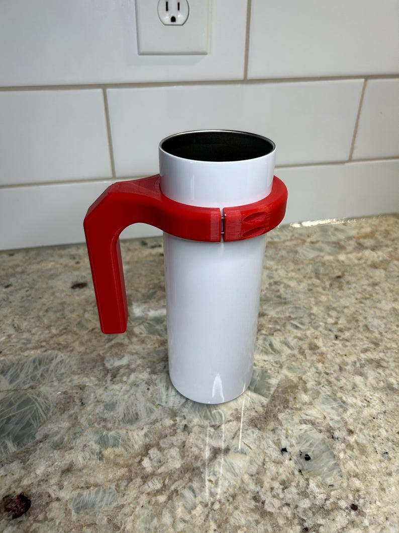 May include: A white tumbler with a red handle. The handle is attached to the tumbler with a clasp. The tumbler is sitting on a granite countertop. The tumbler is designed to hold hot or cold beverages.