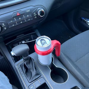 May include: A white can with a red handle and holder sits in a car's cup holder. The car's interior is black and gray, with visible controls and a gear shift. The can appears to be a beverage container.