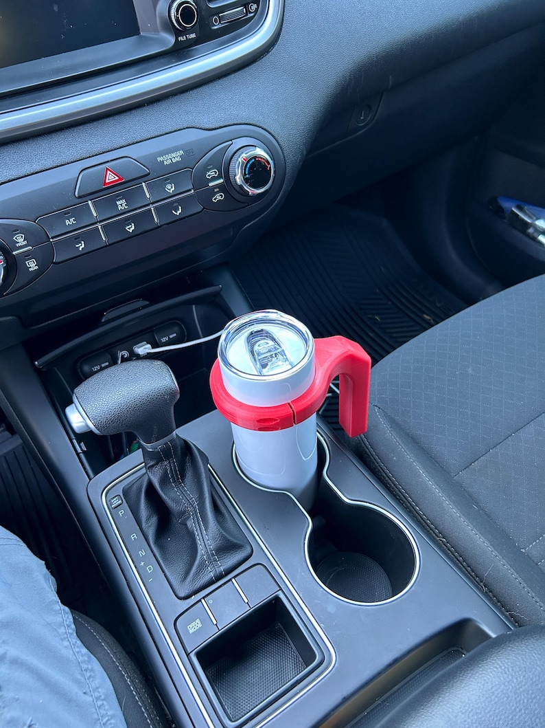 May include: A white can with a red handle and holder sits in a car's cup holder. The car's interior is black and gray, with visible controls and a gear shift. The can appears to be a beverage container.