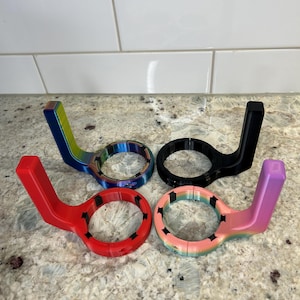 May include: Four colorful, 3D-printed bottle openers. The openers are in red, black, rainbow, and a pink-to-purple gradient. Each has a circular opening with a handle extending from the side. The openers are displayed on a speckled countertop.