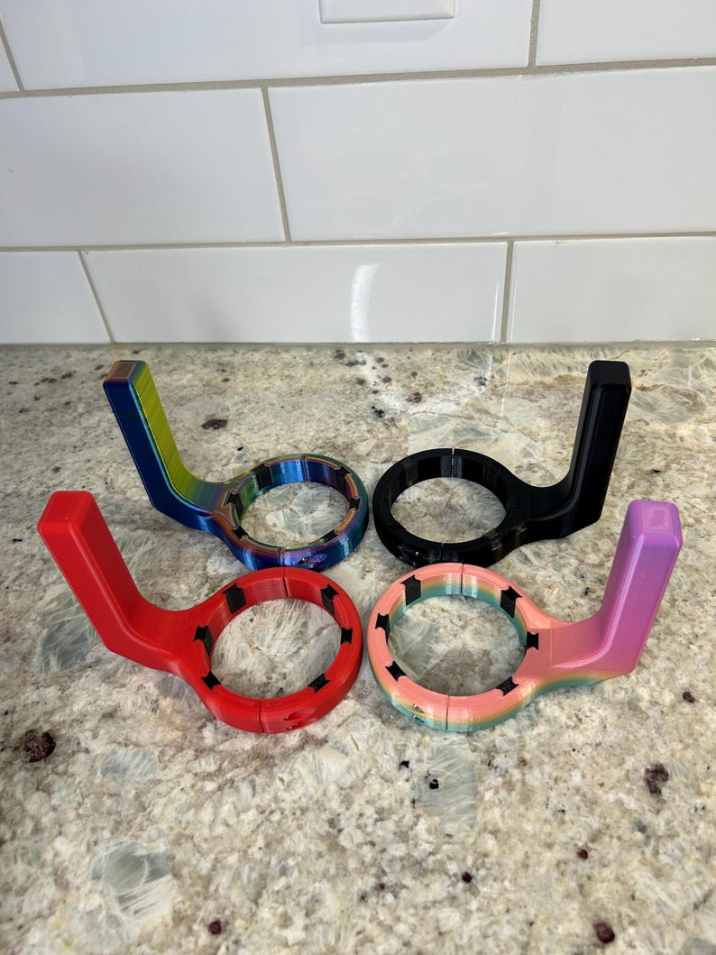 May include: Four colorful, 3D-printed bottle openers. The openers are in red, black, rainbow, and a pink-to-purple gradient. Each has a circular opening with a handle extending from the side. The openers are displayed on a speckled countertop.