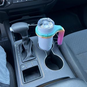 May include: A light purple travel mug with a clear lid and a rainbow-colored handle. The mug is in a car's cup holder, next to the gear shift and other controls.
