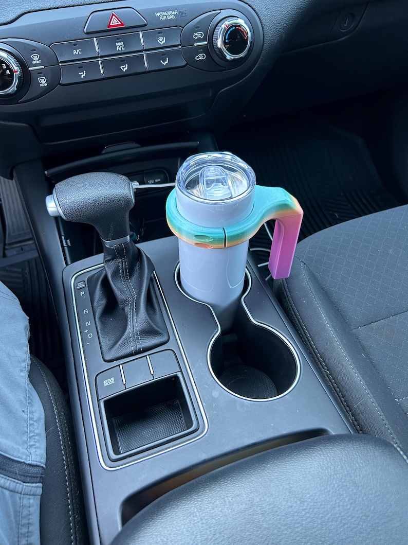 May include: A light purple travel mug with a clear lid and a rainbow-colored handle. The mug is in a car's cup holder, next to the gear shift and other controls.