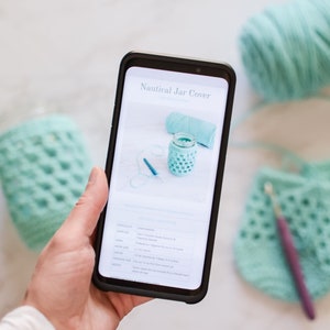 Hand holding a cellphone with the digital crochet pattern shown on it's screen. A crocheting scene shown in the background with blue yarn and crocheted jar covers.