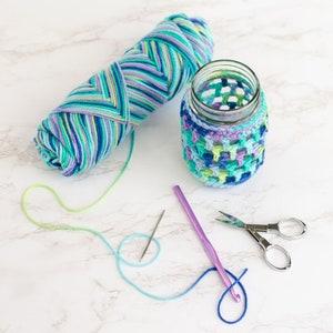 Crocheting scene has a large skein of multi-colored yarn, a mason jar with a finished crochet cozy on it, a purple crochet hook, scissors and a tapestry needle.