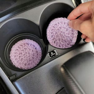 May include: Two lavender crochet car coasters sit in a car's cup holders. A hand is placing one of the coasters into the cup holder. The coasters are round and made of a textured yarn. The car's interior is black.