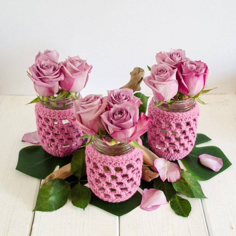 Three glass jar vases decorated with pink crochet covers. Each glass has a small bouquet of pink roses.