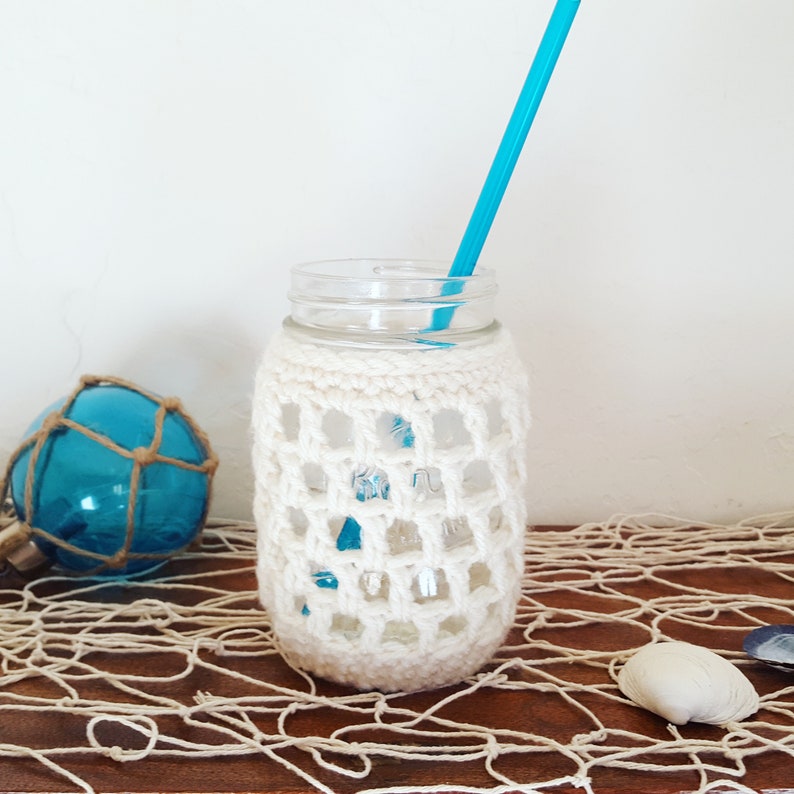 A cream color crochet jar cover shown on a pint size mason jar in a nautical styled scene.