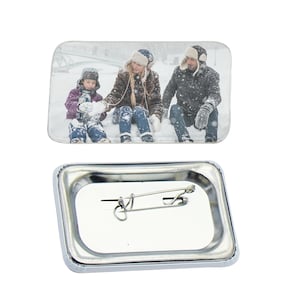May include: A rectangular button with a photo of a family of three in winter clothing playing in the snow. The button has a silver metal backing with a pin.