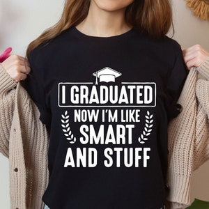 Puede incluir: Camiseta negra con texto blanco que dice "I graduated now I'm like smart and stuff". El texto está rodeado por un gráfico de gorro de graduación y ramas con hojas.