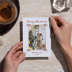 Puede incluir: Una tarjeta de Navidad blanca con un borde rojo y una foto de una familia de pie frente a un &aacute;rbol de Navidad. La tarjeta dice "Feliz Navidad" y "los Daniels, 2021".