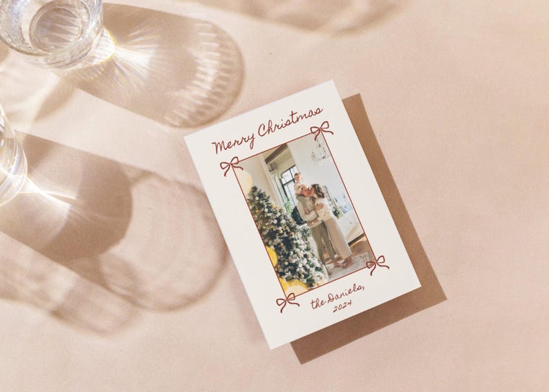 Puede incluir: Una tarjeta de Navidad con una foto de una familia de pie frente a un &aacute;rbol de Navidad decorado. La tarjeta es de color crema con texto rojo que dice "Feliz Navidad" y "the Daniels, 2022".