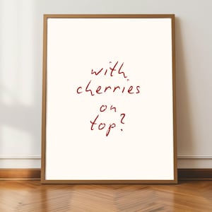 Puede incluir: Un p&oacute;ster enmarcado con la frase "with cherries on top?" en rojo manuscrito sobre un fondo blanquecino. El marco es de color marr&oacute;n claro. El p&oacute;ster se muestra contra una pared blanca y un suelo de madera.